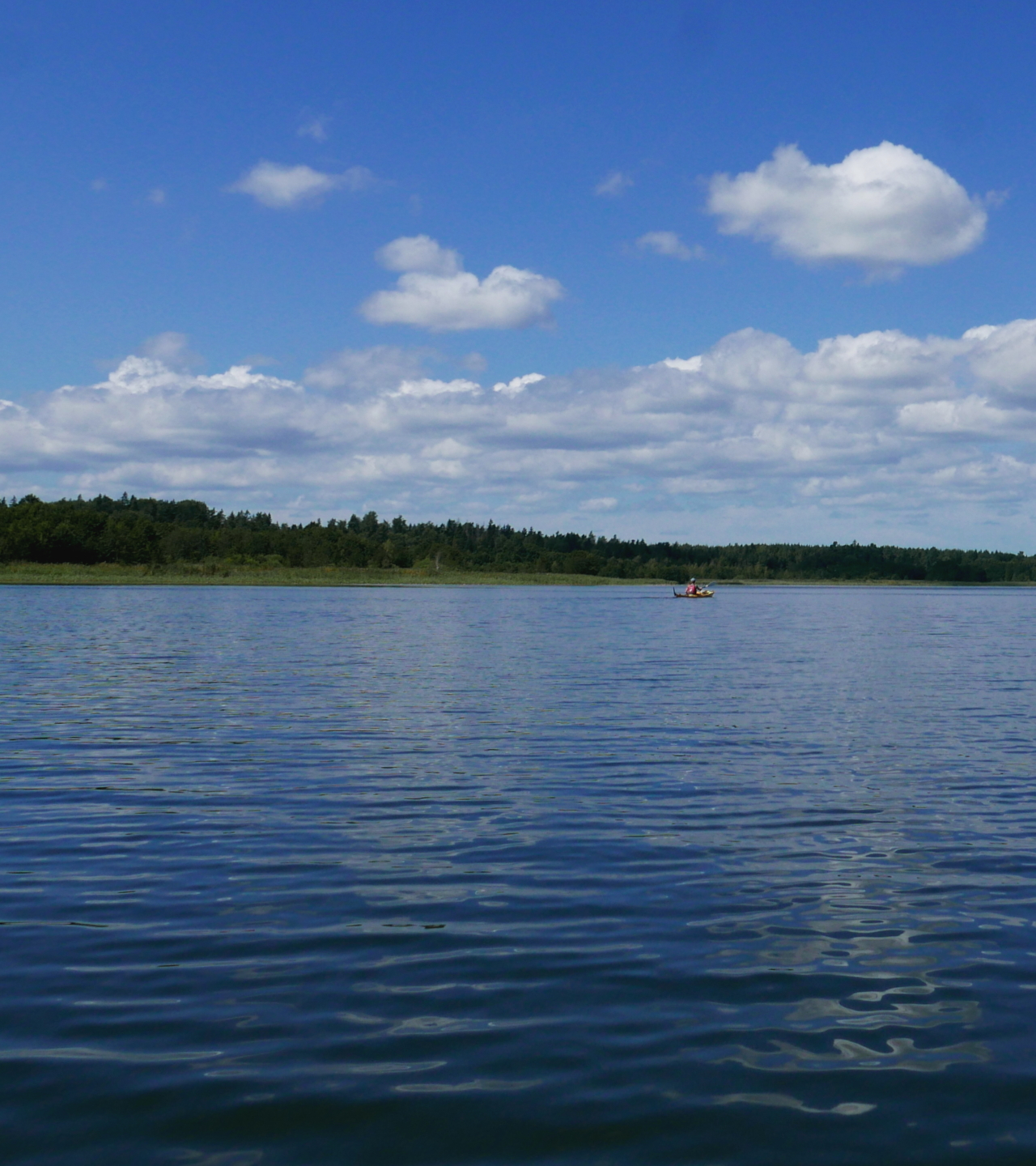 The lower half of the imags shows calm water of a lake, the upper half mostly sky partly covered by gray but fluffy clouds. A stripe of land cuts the middle and a kayaker can be seen to the right.