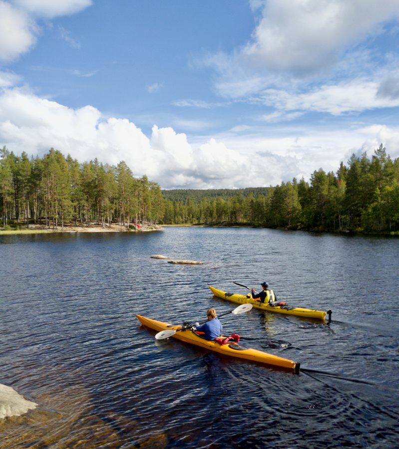 Two people in kayaks, one yellow and one orange, paddle across a lake. It's a nice summer's day with a blue sky and a few white cumulus clouds. Original photo by Christer Gundersen.