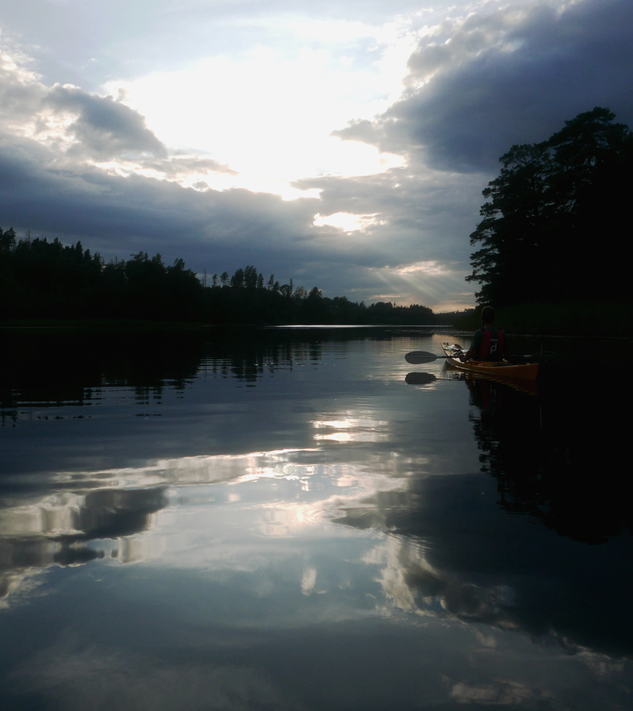 The sun setting through clouds reflects in almost perfectly still water. The lake narrows off towards forests in the distance and in the dark reflection of a tree it is possible to make out the shape of a person sitting still in a kayak.
