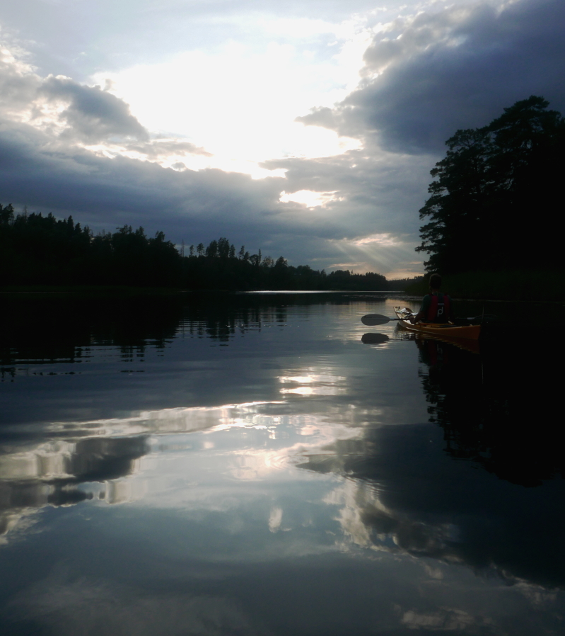 The sun setting through clouds reflects in almost perfectly still water. The lake narrows off towards forests in the distance and in the dark reflection of a tree it is possible to make out the shape of a person sitting still in a kayak.