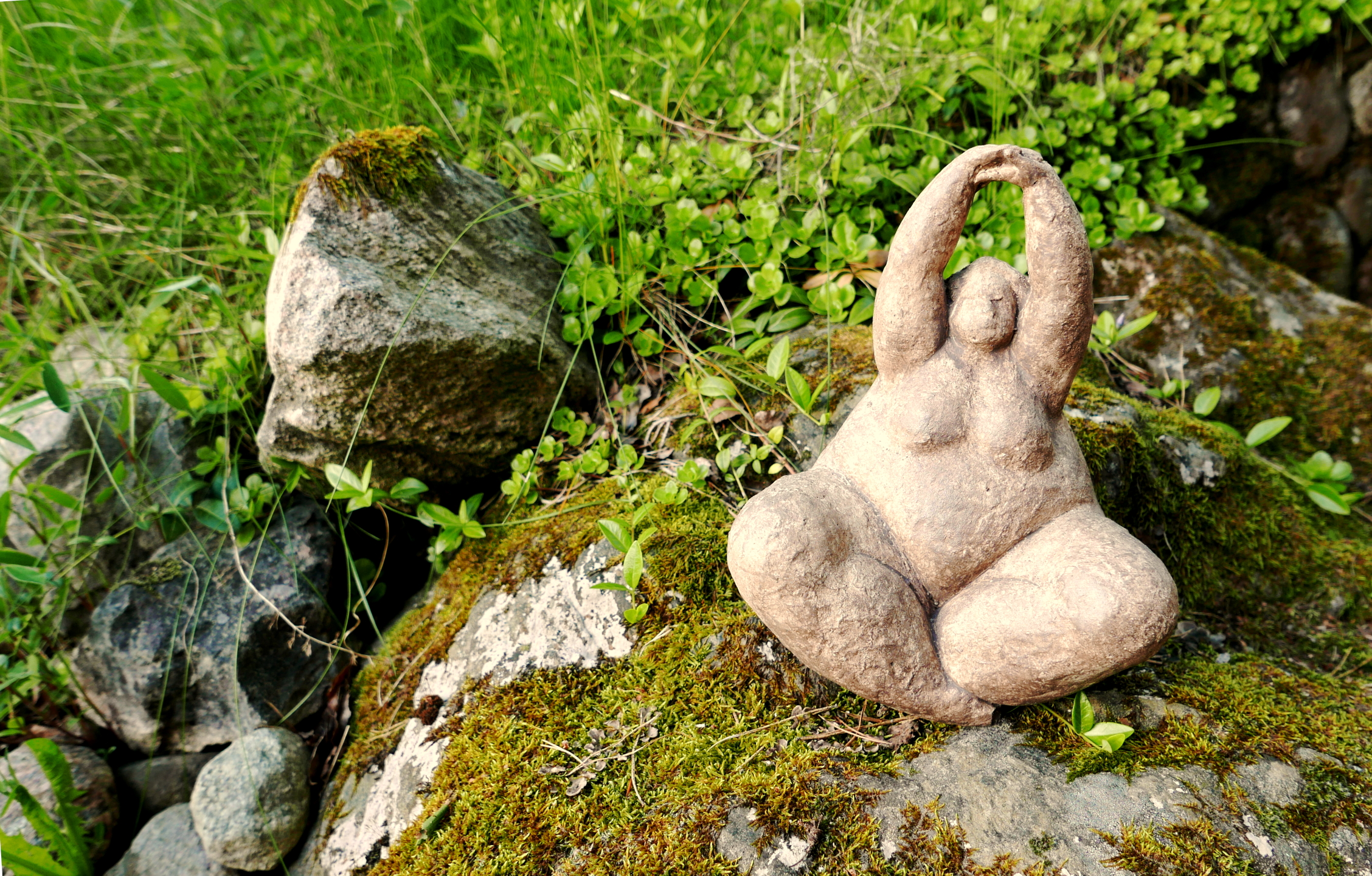 A stone statue, similar in shape to a Venus figurine, sits in a yoga position on a moss covered stone with leaves and grass in the background.