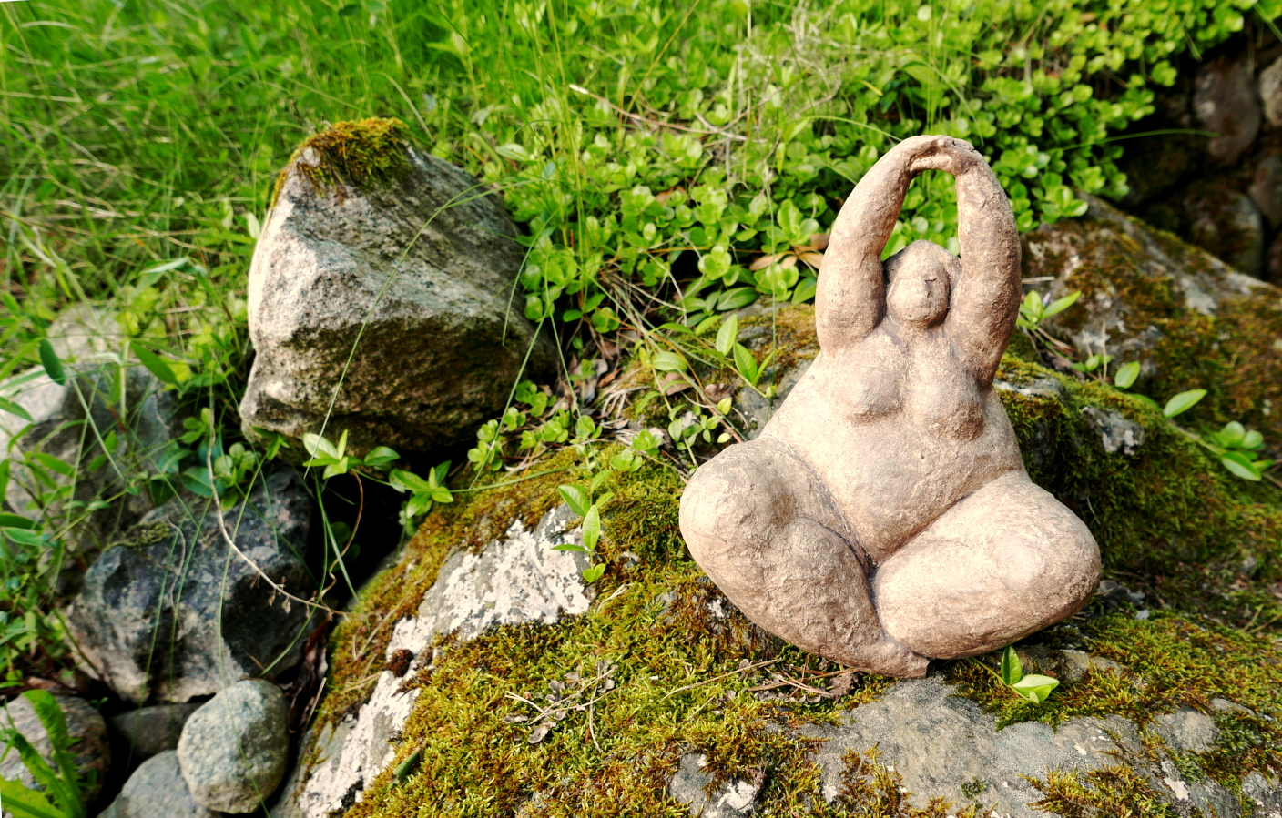 A stone statue, similar in shape to a Venus figurine, sits in a yoga position on a moss covered stone with leaves and grass in the background.
