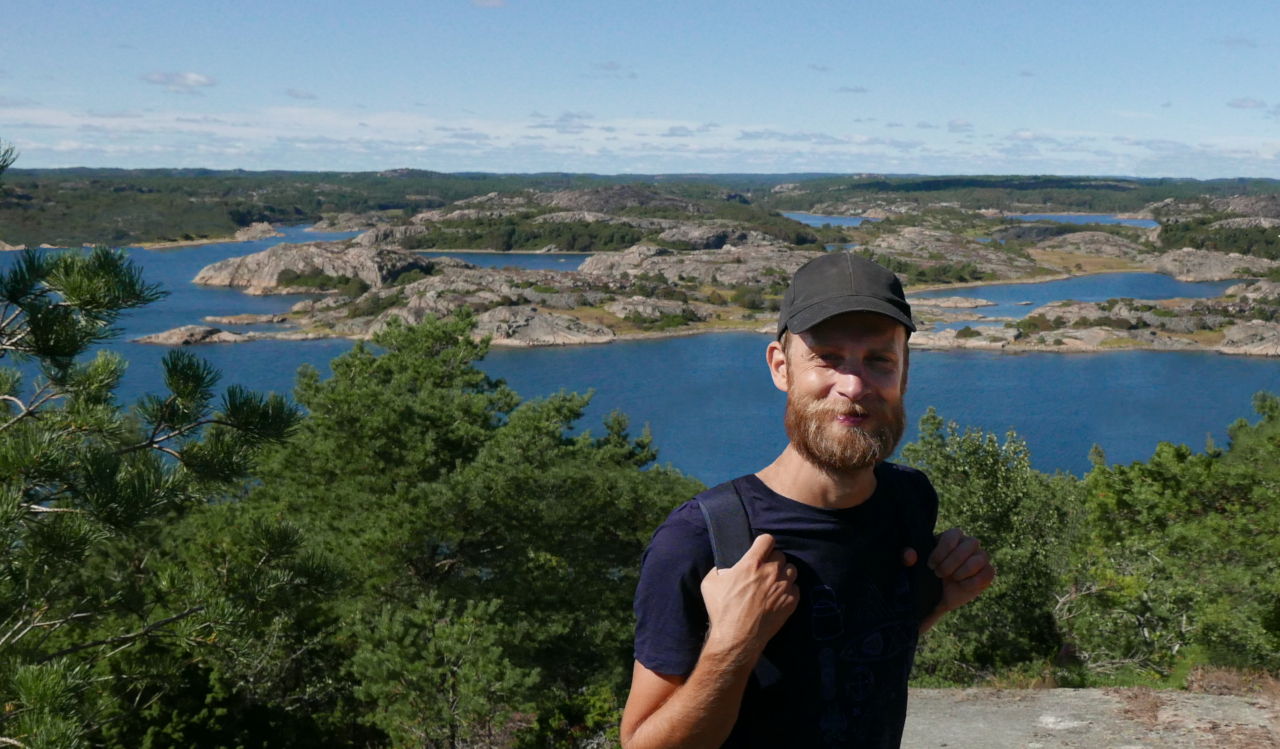 An image of Tino smiling at the camera while standing on a hill in front of water, cliffs and forest. There are bushes right behind him and the sun is shining bright from a light blue sky.
