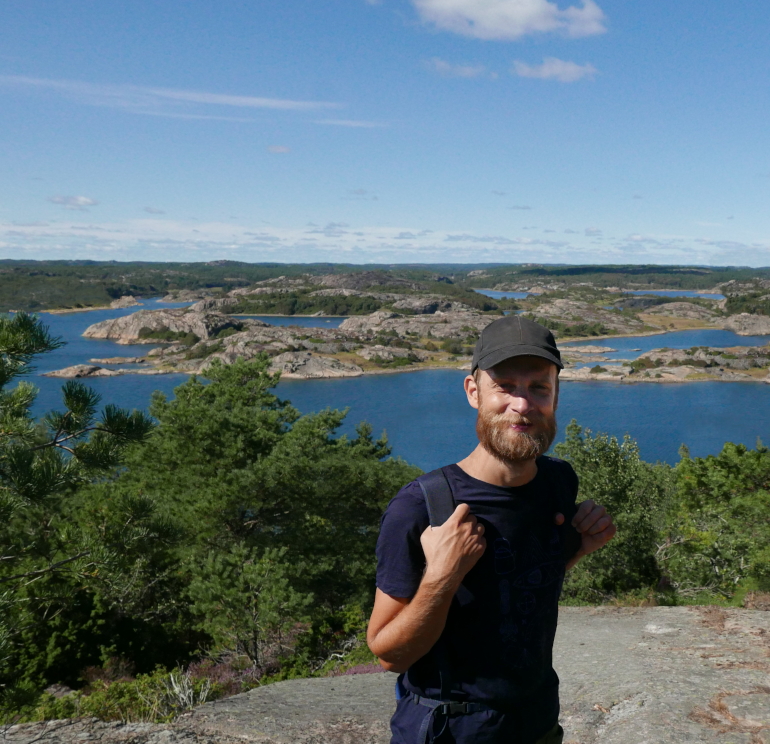 An image of Tino smiling at the camera while standing on a hill in front of water, cliffs and forest. There are bushes right behind him and the sun is shining bright from a light blue sky.
