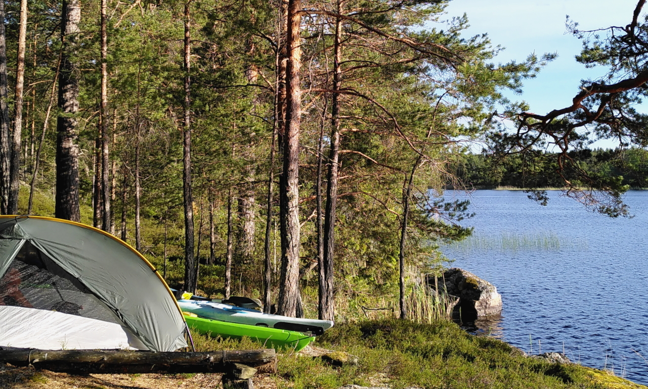 A tent stands on the edge of the woods, next to calm water. Two kayaks lie slightly hidden behind the tent. It is a sunny day.