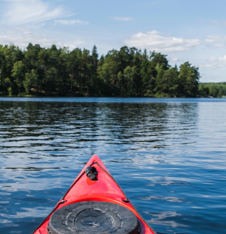 The tip of a red kayak is seen in the foreground, on a calm lake with trees and a summer's sky in the distance. Photo by Oliver Schmid.