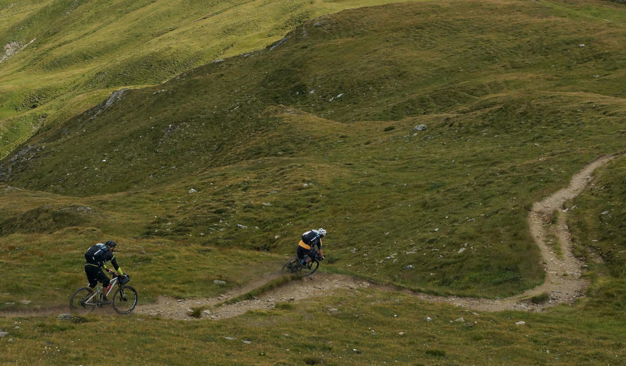 Two people seen from a distance cycling down a mountain path. The terrain is grass and rocks, and the image gives an impression of speed. Original photo by Sebastian Marx.