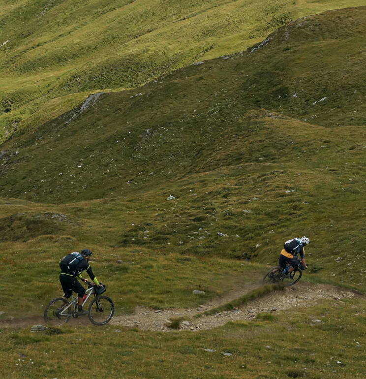 Two people seen from a distance cycling down a mountain path. The terrain is grass and rocks, and the image gives an impression of speed. Original photo by Sebastian Marx.
