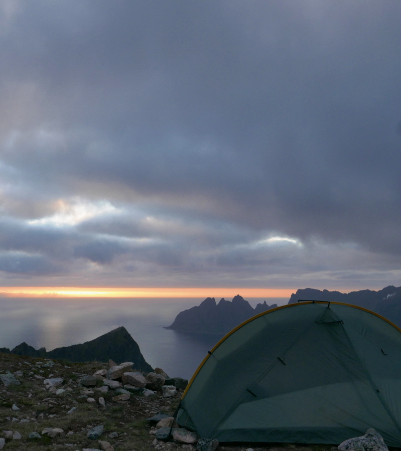 A tent stands in the foreground on a cliff overlooking a fjord, a steep peninsula and the ocean. It is midnight and the sun that does not set can be seen towards the horizon.