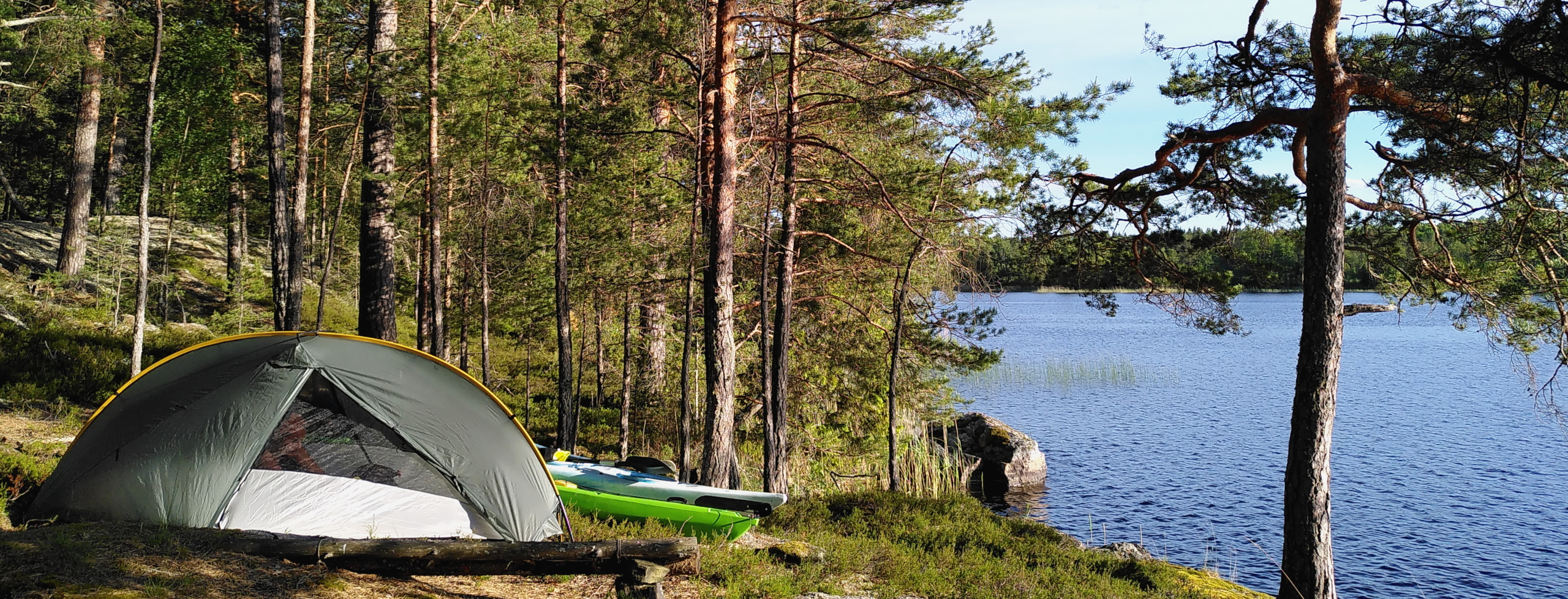 A tent stands on the edge of the woods, next to calm water. Two kayaks lie slightly hidden behind the tent. It is a sunny day.