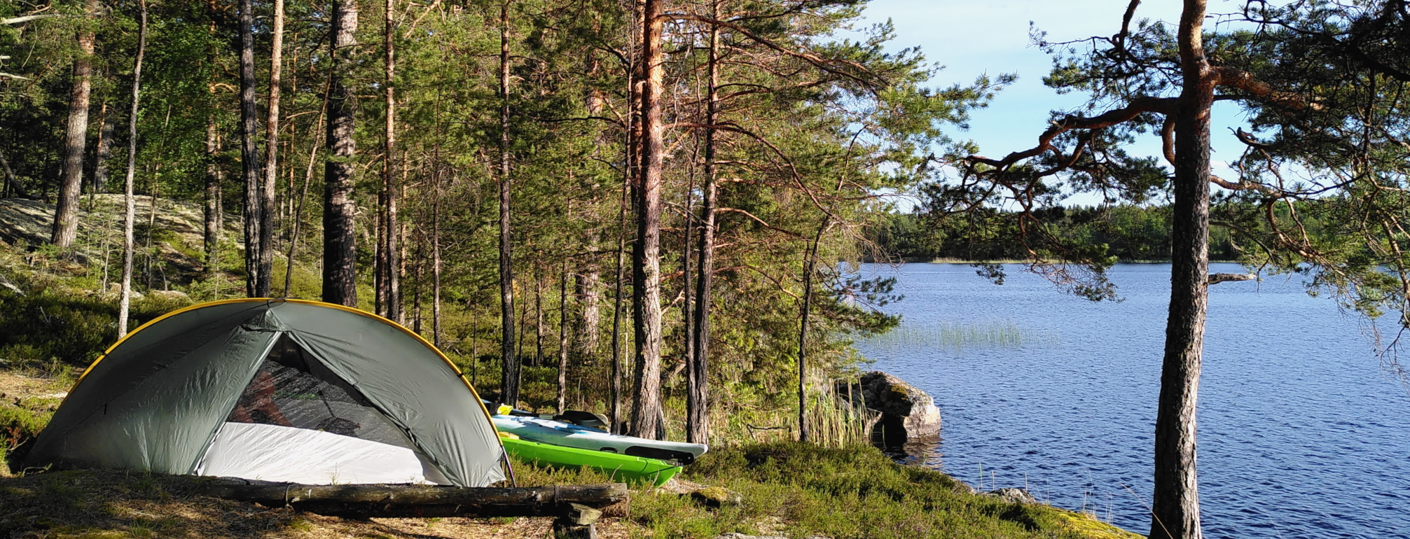 A tent stands on the edge of the woods, next to calm water. Two kayaks lie slightly hidden behind the tent. It is a sunny day.
