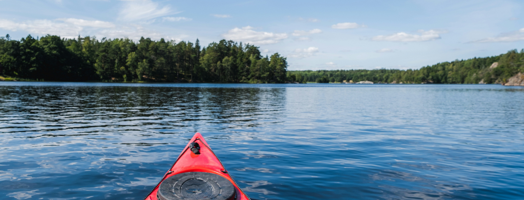 The tip of a red kayak is seen in the foreground, on a calm lake with trees and a summer's sky in the distance. Photo by Oliver Schmid.